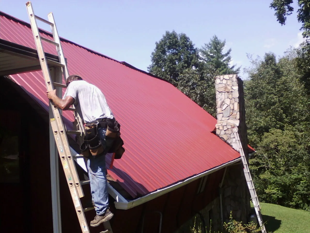Roofer on a ladder beside a red metal roof and stone chimney — Soffit & Fascia Repair by Roofing Brothers in Coltons Point